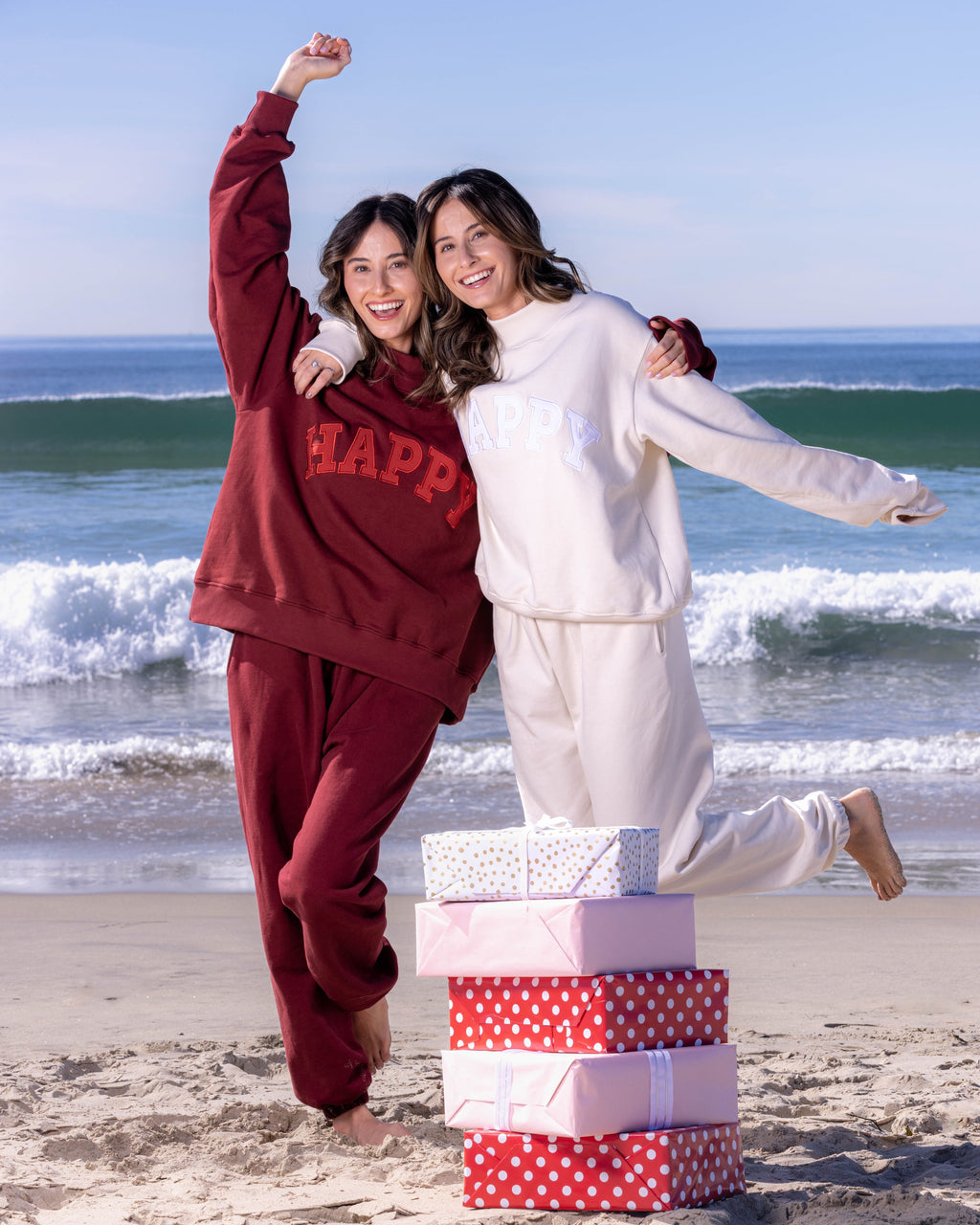Two people standing on a beach with stacked gift boxes, one wearing a red 'HAPPY' sweatshirt.