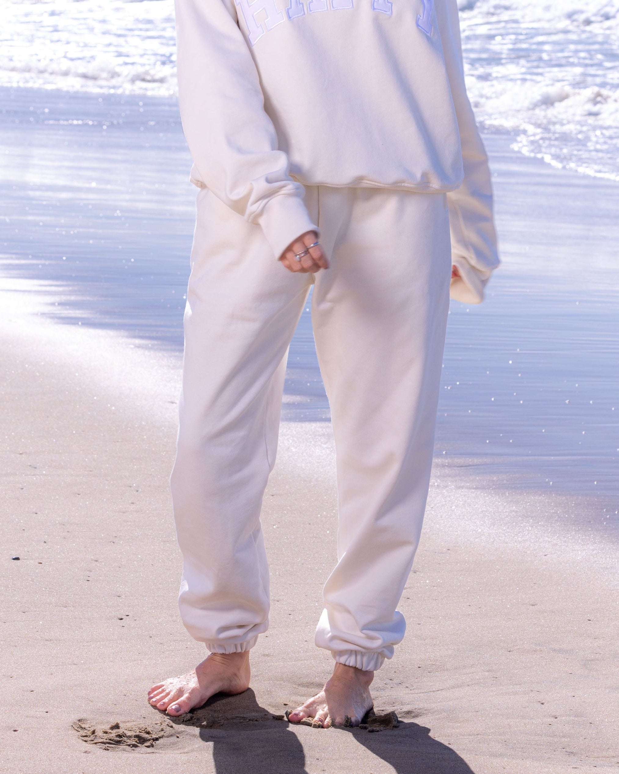 Person wearing a white matching sweat set standing on a beach with ocean waves in the background