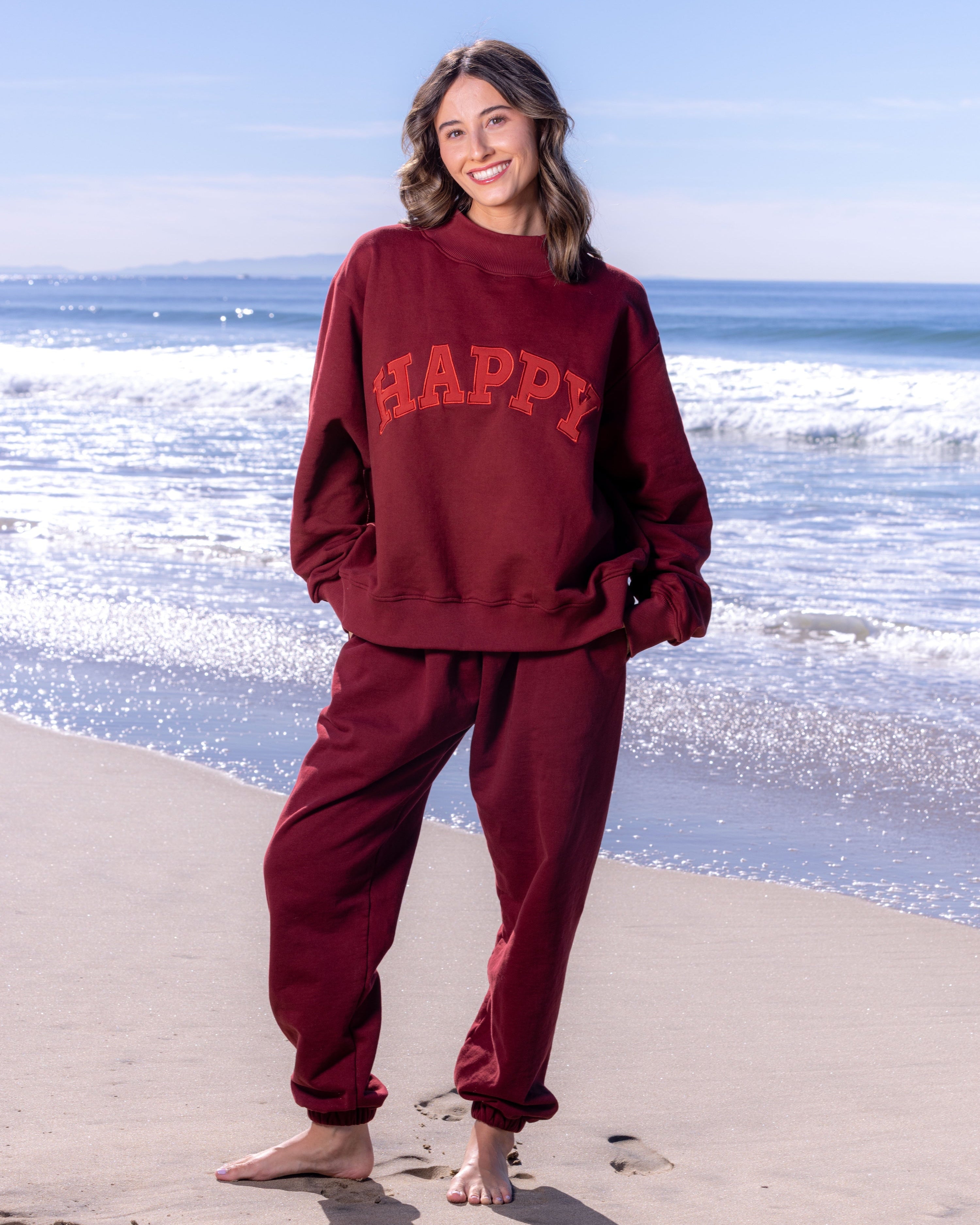 Person wearing a maroon 'HAPPY' sweatshirt and pants on a beach.