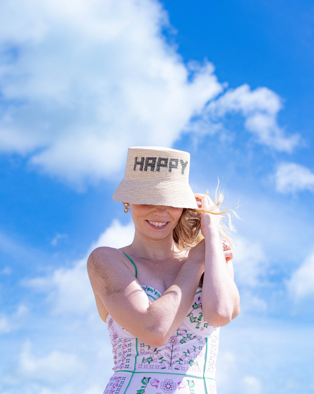 Woman wearing a floral swimsuit and straw hat with 'HAPPY' text against a blue sky.
