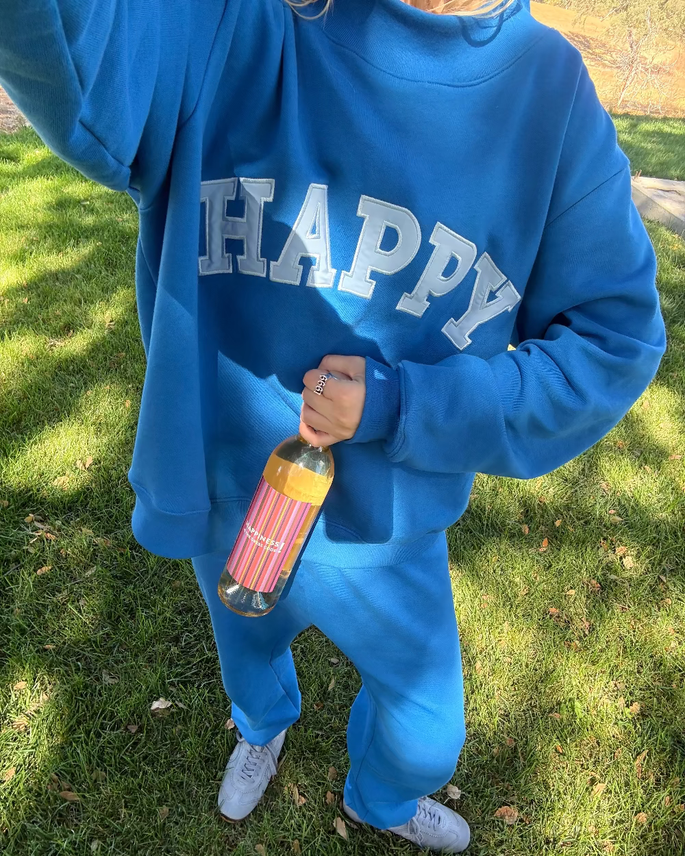 Person wearing a blue 'HAPPY' sweatshirt holding a colorful bottle on grass.