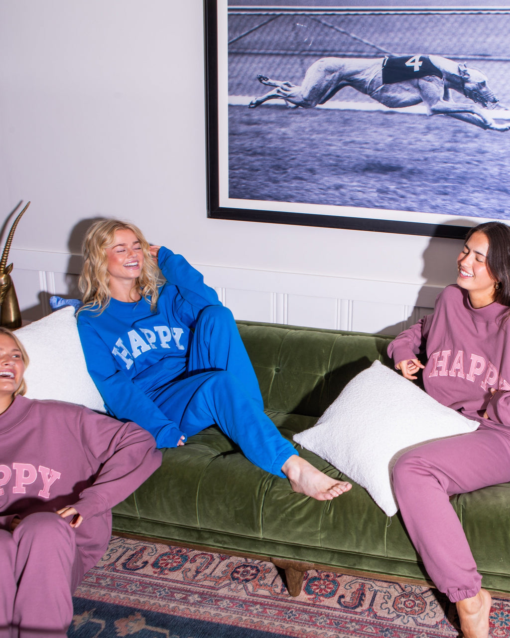 Two women sitting on a green couch wearing matching 'HAPPY' sweatshirts and pants in a living room.